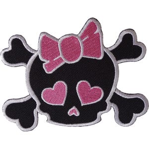 Pode incluir: Um emblema bordado apresentando um crânio preto com um laço rosa no topo e dois olhos em formato de coração rosa. O crânio está posicionado à frente de dois ossos pretos cruzados, tudo contornado por uma borda branca.