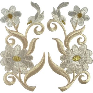 Könnte beinhalten: Zwei symmetrische Stickaufnäher mit weißen Blüten, goldenen Mitten sowie beigefarbenen, geschwungenen Stielen und Blättern. Das florale Design besteht aus einer großen unteren und einer kleineren oberen Blüte mit detaillierter, strukturierter Stickerei.