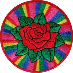 Puede incluir: Un parche bordado circular que presenta una rosa roja central con hojas verdes. El fondo consiste en rayas vibrantes y radiantes en colores del arcoíris, incluyendo rosa, amarillo, verde y azul, todo encerrado por un borde rojo cosido.