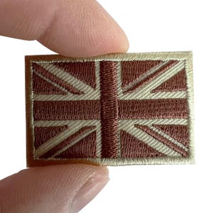 May include: A rectangular embroidered patch featuring the Union Jack flag design. The pattern consists of a central vertical stripe and diagonal lines in a deep brown thread, contrasted against a light beige background with a matching border.