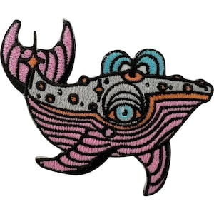 May include: An embroidered patch shaped like a stylised whale with a large central eye. The design features a light grey upper body with dark spots, a pink striped underbelly, and bright blue accents on the dorsal fin and eye, all outlined in black.