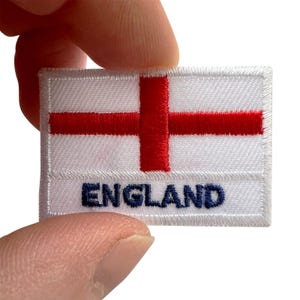 May include: A rectangular embroidered patch featuring the flag of England, which consists of a white background with a centred red cross. The word ENGLAND is stitched in dark blue capital letters along the bottom portion of the white fabric.