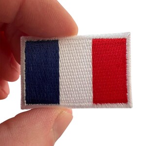 May include: A rectangular embroidered patch featuring the French national flag design with vertical stripes in navy blue, white, and red, finished with a white stitched border around the entire perimeter of the fabric emblem.