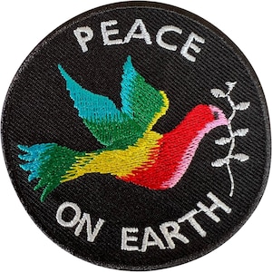 May include: A circular black embroidered patch featuring a colourful bird in flight with teal, yellow, and red feathers. The bird holds a white olive branch in its beak. The white text PEACE is arched above the bird, and ON EARTH is below.