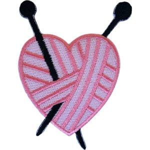May include: An embroidered patch shaped like a heart made of light pink yarn. Two black knitting needles are crossed diagonally through the centre of the heart. The design features detailed stitching that mimics the texture of wound yarn.