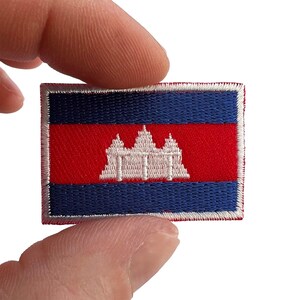 May include: A rectangular embroidered patch featuring the flag of Cambodia. The design consists of a central red horizontal band with a white depiction of the Angkor Wat temple, flanked by two blue horizontal bands, all bordered by white stitching.