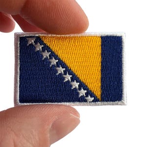 May include: A rectangular embroidered patch featuring the flag of Bosnia and Herzegovina. The design includes a dark blue field with a yellow triangle and a diagonal row of nine white five-pointed stars, all bordered by a white stitched edge.