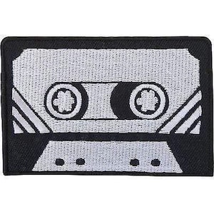 Può includere: Una toppa ricamata rettangolare con un design in bianco e nero di una cassetta vintage. La toppa mostra la forma iconica di una cassetta con due bobine circolari, un'area centrale per l'etichetta e linee inclinate sui lati.