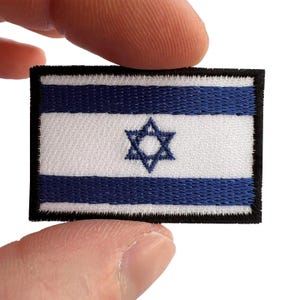 Puede incluir: Un parche rectangular bordado que muestra la bandera de Israel. El diseño consiste en un fondo blanco con dos franjas horizontales azules y una estrella de David azul en el centro. El parche tiene un borde grueso cosido en color negro.