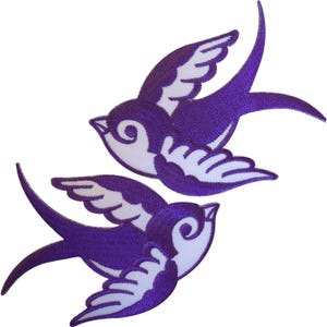 Könnte beinhalten: Zwei gestickte Schwalben-Aufnäher in einem violett-weißen Farbschema. Jeder Vogel ist in einer Flugposition mit ausgebreiteten Flügeln dargestellt, wobei detaillierte Stickmuster die Federn und die Körperform der Vögel deutlich hervorheben.