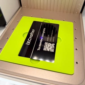 Puede incluir: Una tarjeta de presentación rectangular negra con texto blanco que dice iXCrafter, Tu nombre completo y un código QR, colocada dentro de una plantilla de alineación cuadrada de color verde lima brillante sobre una base de herramienta beige con marcas.
