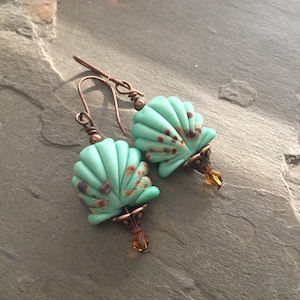 May include: A pair of turquoise blue shell-shaped earrings with copper wire and brown accents. The earrings have a copper hook and a small amber-colored crystal at the bottom.