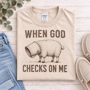 Puede incluir: Una camiseta beige de manga corta con un gráfico marrón de una oveja con un cubo de metal sobre la cabeza. El texto sobre la oveja dice "WHEN GOD" y el texto debajo dice "CHECKS ON ME" en una fuente marrón de estilo bloque desgastado.