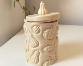 May include: A cylindrical beige container with a matching lid featuring a small pumpkin-shaped handle. The exterior surface is decorated with a repeating embossed pattern of various pumpkin and gourd shapes in a uniform matte beige finish.