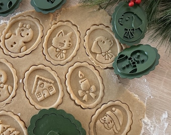 May include: A set of dark green oval cookie stamps with various embossed designs including a cat, dog, house, candle, and Santa Claus. The stamps are shown pressing into rolled dough, creating detailed impressions of the festive holiday icons.