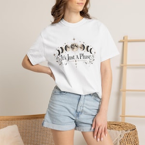 Könnte beinhalten: Ein weißes Kurzarm-T-Shirt mit einer Grafik von Mondphasen, die in einem Bogen über dem Text It's Just A Phase angeordnet sind. Das Design enthält Sterne und botanische Elemente in Braun-, Schwarz- und Beigetönen auf der Brust.