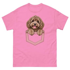 Può includere: Una t-shirt rosa brillante a maniche corte con una stampa grafica di un cucciolo marrone dal pelo riccio che spunta da una finta tasca. Il cucciolo ha orecchie pendenti, naso nero e bocca aperta con la lingua visibile, appoggiando le zampe.