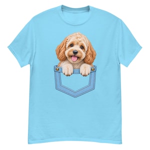 Peut inclure: Un t-shirt bleu clair à manches courtes avec une impression graphique représentant un chiot beige aux poils bouclés qui sort d'une poche en jean bleue. Le chiot a des oreilles tombantes, un nez noir et la gueule ouverte avec la langue visible.