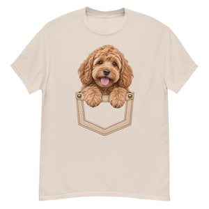Può includere: Una t-shirt a maniche corte color beige chiaro con una stampa grafica di un cucciolo marrone dorato, soffice e riccio, che spunta da una finta tasca sul petto. Il cucciolo ha grandi occhi scuri, un naso nero e la lingua visibile.