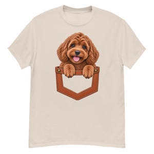 Può includere: Una t-shirt beige chiaro con una stampa grafica di un cucciolo marrone soffice dal pelo riccio con la lingua rosa di fuori, che sembra spuntare da una tasca in stile pelle marrone con cuciture visibili e dettagli di rivetti metallici.