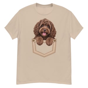 Può includere: Una t-shirt a maniche corte color beige chiaro con una stampa grafica di un cucciolo marrone dal pelo riccio che spunta da una finta tasca sul petto. Il cucciolo ha occhi scuri, una lingua rosa e le zampe anteriori sul bordo della tasca.