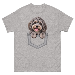 Puede incluir: Una camiseta gris jaspeado de manga corta con un estampado gráfico de un cachorro de pelo rizado color marrón y crema asomándose por un bolsillo de mezclilla falso. El cachorro tiene ojos oscuros, nariz negra y lengua rosa visible.