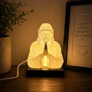 May include: A white, translucent, illuminated lamp shaped as a bust of Jesus with hands pressed together in prayer, wearing a crown of thorns. The lamp sits on a rectangular black base and emits a warm, soft glow from within the figure.