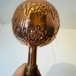 May include: A spherical copper object with an embossed floral pattern featuring fleur-de-lis motifs. The copper sphere is attached to a cylindrical copper neck that connects to a dark, ribbed wooden handle, creating a tool-like implement.