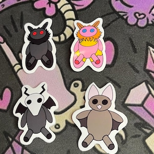 Könnte beinhalten: Vier gestanzte Aufkleber mit Cartoon-Kreaturen: ein schwarzes flauschiges Wesen mit roten Knopfaugen, ein rosa-orangefarbenes flauschiges Wesen mit blauen Knopfaugen, ein graues Wesen mit weißem Totenkopf-Kopf und eine beige Fledermaus.