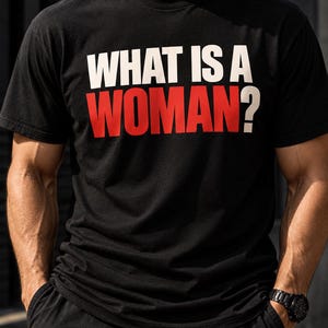 May include: A black short-sleeved t-shirt featuring the text "WHAT IS A WOMAN?" printed across the chest. The word "WHAT IS A" is in white capital letters, while the word "WOMAN?" is in large, bold red capital letters.