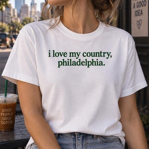 Könnte beinhalten: Ein weißes kurzärmeliges T-Shirt mit Rundhalsausschnitt und dem dunkelgrünen Schriftzug "i love my country, philadelphia." in einer einfachen, kleingeschriebenen serifenlosen Schriftart auf der Brust, aus Standard-Baumwollstoff.