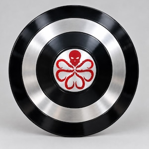 Puede incluir: Un escudo circular con anillos concéntricos alternos de metal negro brillante y plata cepillada. El centro del escudo muestra un gráfico rojo de una calavera con seis apéndices en forma de tentáculos que se extienden hacia abajo.