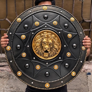 May include: A circular shield featuring a central gold-coloured lion head relief. The shield is primarily black with a weathered finish, accented by gold-coloured circular studs and smaller silver-coloured rivets arranged in a star-like pattern.