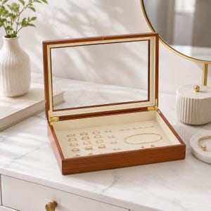 May include: A rectangular wooden jewelry box with a clear glass hinged lid and a cream-colored interior lining. The box contains several gold rings, small stud earrings, a delicate gold chain bracelet, and a thin gold bangle arranged inside.