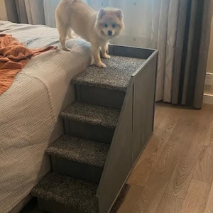 May include: A small, light-colored dog stands on a gray pet step. The step has three carpeted stairs and a gray wooden frame. The dog is looking at the camera. The step is placed next to a bed.