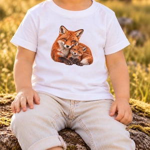 May include: A white short-sleeved t-shirt featuring a detailed graphic print of an adult red fox lying down with a small fox kit nestled against its side. The fox illustration displays realistic orange, white, and black fur textures.