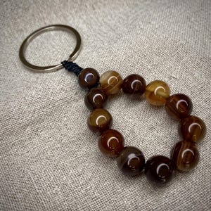 May include: A keychain featuring a circular metal ring attached to a teardrop-shaped loop of twelve polished, spherical brown and tan banded agate beads. The beads are strung on a dark cord, showcasing natural variations in color and translucent banding.