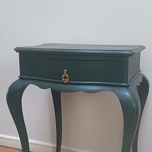 May include: A dark green side table featuring a single drawer with a decorative gold-toned metal pull. The table has a rectangular top with a scalloped apron and four curved cabriole legs that extend outward from the base of the drawer unit.