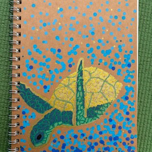 May include: A spiral-bound notebook with a tan cover featuring a painted sea turtle. The turtle has a textured yellow shell and dark green patterned flippers. The background is decorated with numerous blue and light blue dots of varying sizes.