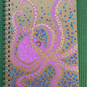 Può includere: Un taccuino a spirale con una copertina color marrone chiaro che presenta un polpo rosa dipinto a mano. Il polpo ha ventose gialle sui tentacoli e un occhio giallo. Lo sfondo è decorato con punti dipinti in viola e blu brillante.