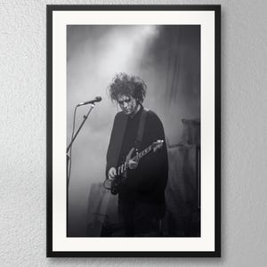 May include: A black and white framed photograph of a musician with messy hair performing on stage. The individual wears a dark long-sleeved shirt and plays an electric guitar while standing in front of a microphone stand under bright stage lights.