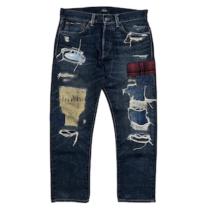 Può includere: Jeans in denim scuro con finitura usurata, strappi multipli e bordi sfilacciati. I pantaloni presentano una toppa in tessuto scozzese rosso e nero, una toppa rettangolare beige con testo scuro e una chiusura con patta a bottoni.