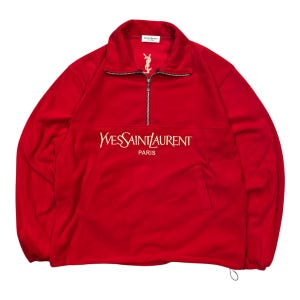 Puede incluir: Un suéter rojo brillante de manga larga con cierre de cremallera en el cuello. El pecho muestra el texto bordado en blanco Yves Saint Laurent Paris, con un logotipo más pequeño a juego cerca del cuello y una etiqueta de marca blanca interior.