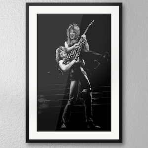 May include: A black and white framed photograph depicting a musician with long hair holding a guitar while being lifted by another long-haired individual on a stage. The scene captures a high-energy performance with both figures wearing dark clothing.