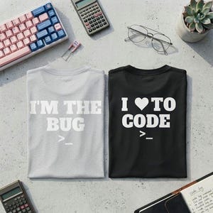 May include: A light gray t-shirt with the white text I'M THE BUG >_ and a black t-shirt with the white text I (heart symbol) TO CODE >_. Both shirts feature short sleeves and a crew neckline, displayed flat on a textured surface with office items.