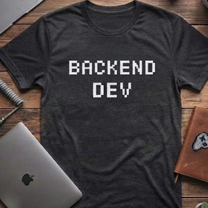 May include: A dark charcoal gray short-sleeve t-shirt featuring the text BACKEND DEV printed in a white, pixelated, blocky font across the center of the chest. The shirt has a classic crew neckline and a standard fit with a heathered texture.