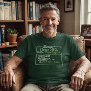 May include: A dark green short-sleeved t-shirt featuring a light green graphic of a computer terminal screen with the text I Speak COBOL Better Than English and various lines of COBOL programming code printed on the front chest area.