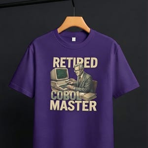 Puede incluir: Una camiseta morada de manga corta con un estampado gráfico de una persona con traje escribiendo en un ordenador antiguo. El texto encima y debajo de la ilustración dice RETIRED COBOL MASTER en letras mayúsculas grandes y claras.