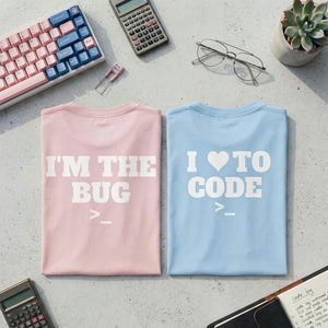 Może przedstawiać: Jasnoróżowy t-shirt z białym napisem I'M THE BUG oraz jasnoniebieski t-shirt z białym napisem I HEART TO CODE, oba z symbolem znaku zachęty terminala. Koszulki leżą na biurku obok klawiatury, kalkulatora i sukulenta w doniczce.