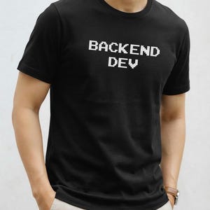 May include: A black short-sleeved t-shirt featuring the white pixelated text "BACKEND DEV" printed across the chest. The garment has a classic crew neckline and a straight hem, providing a simple and clean look for everyday casual wear.